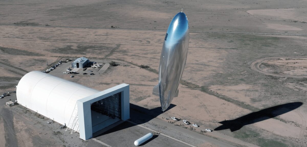 A large silver rocket prototype stands vertically on a desert test site near a hangar.