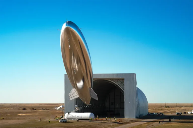 Silver airship emerging from large hangar.